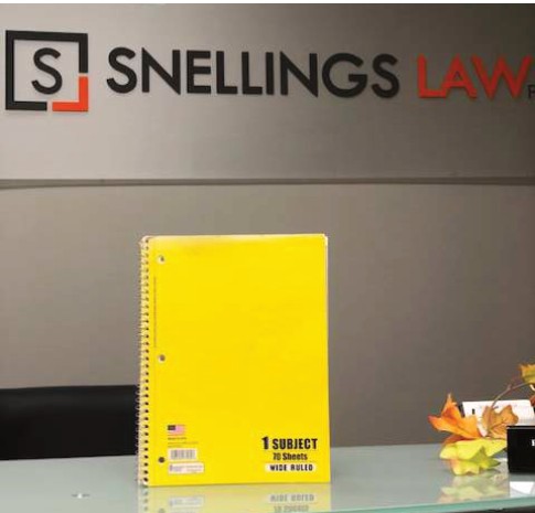 A yellow spiral notebook standing on a desk in front of the Snellings Law sign
