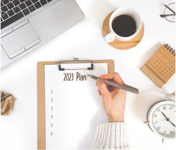 A hand writing a 2023 plan on a clipboard beside a laptop, coffee and an alarm clock