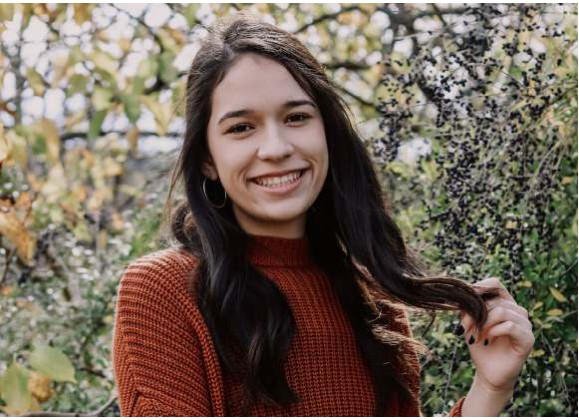 A young woman with long dark hair smiling outdoors in a rust-colored sweater