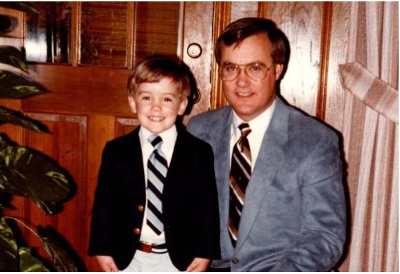 A vintage photo of a young boy in a suit and tie standing with his father