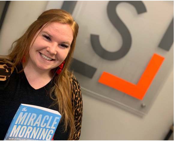A smiling woman holding a copy of The Miracle Morning in front of the Snellings Law logo